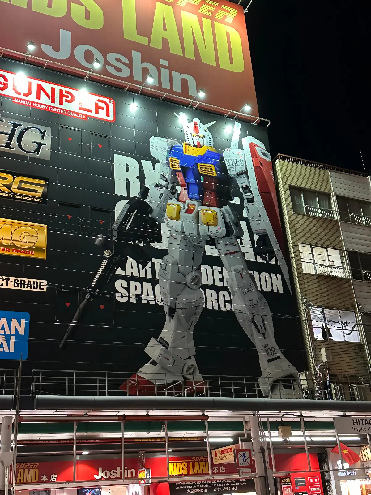 A brightly lit building features a large mural of a Gundam robot from the anime series, surrounded by colorful advertisements and signs in Japanese at night.