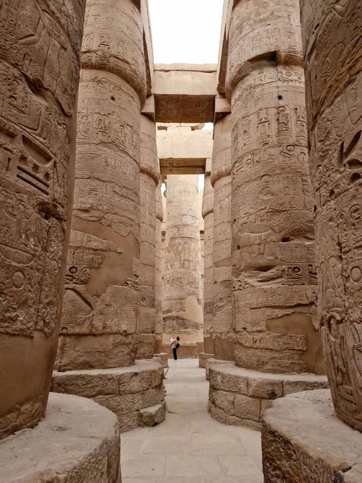 Ancient stone columns with intricate carvings, towering in a row, forming part of a historic temple. The pillars are massive and weathered, creating a narrow pathway between them. A person stands at the far end, highlighting their scale.