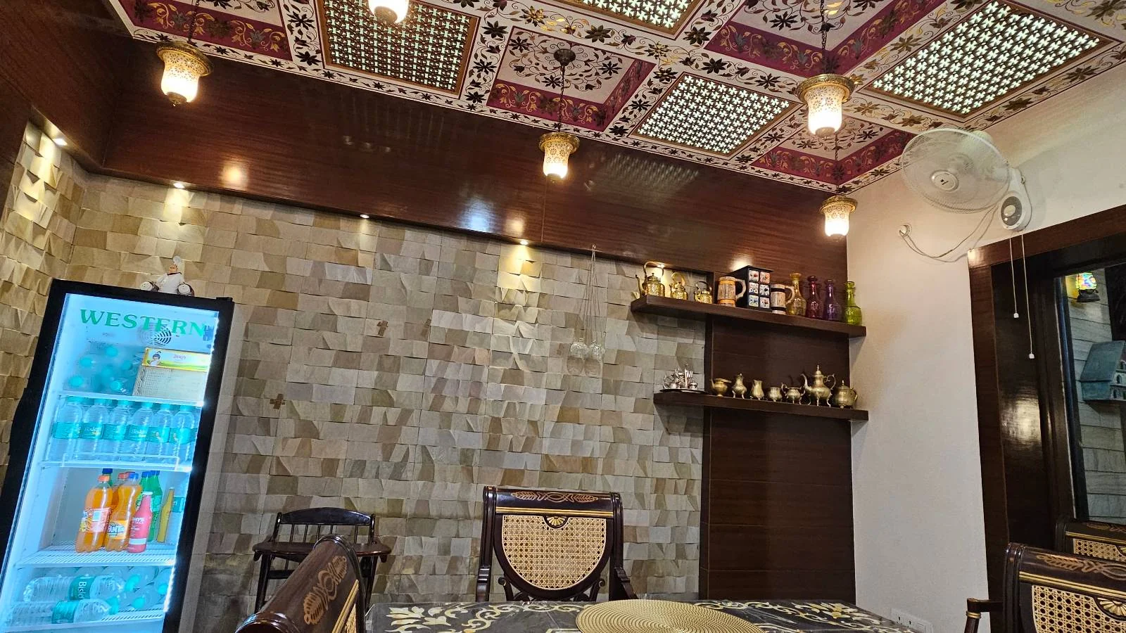 A cozy room with mosaic ceiling lights, a wall-mounted shelf with decorations, a light stone wall, a glass-front refrigerator with drinks, and wooden furniture. A wall fan and framed mirror are also visible.