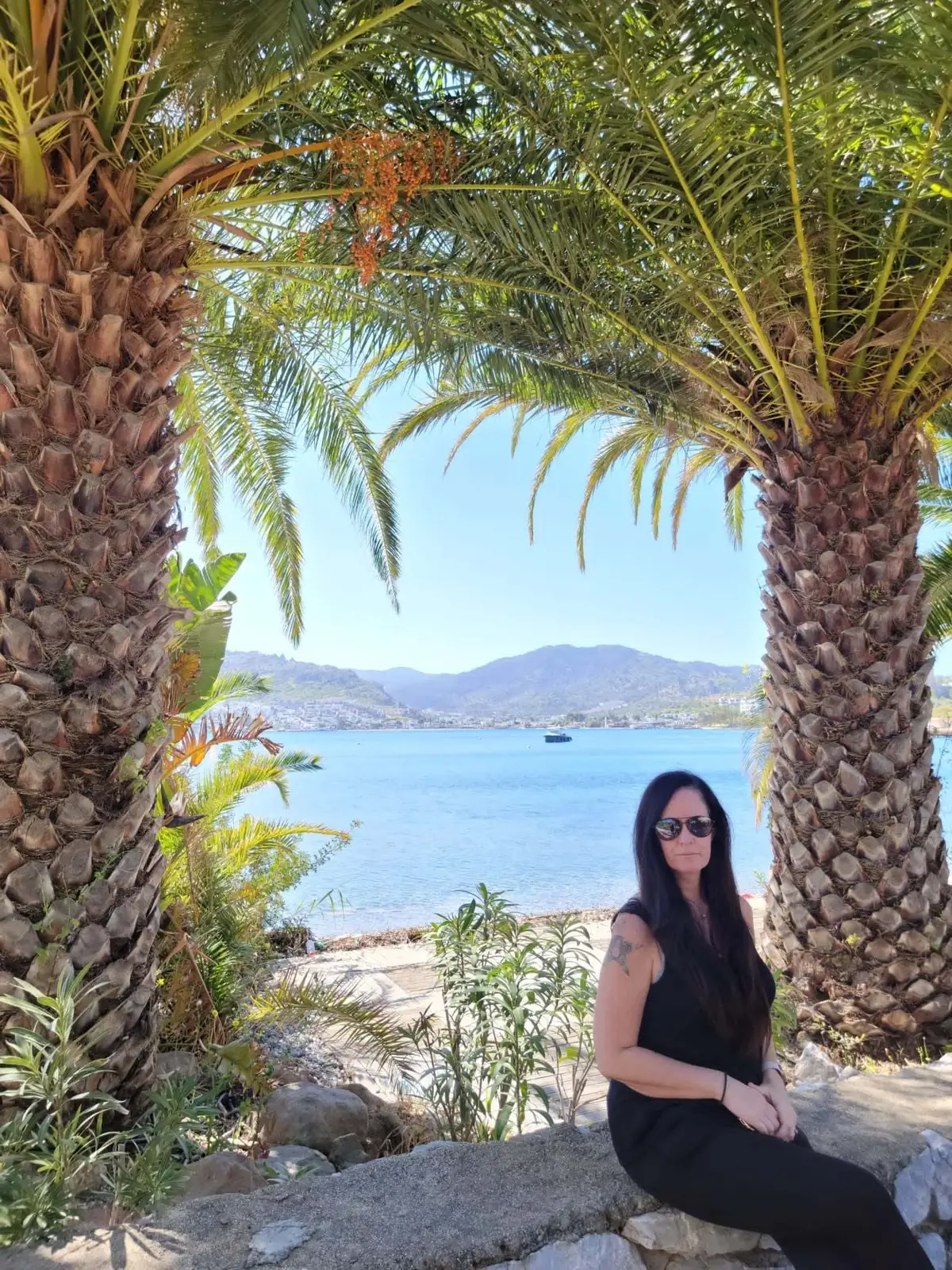A woman in black clothing sits on a stone bench between two palm trees, with a scenic view of a blue lake and distant mountains under a clear sky.