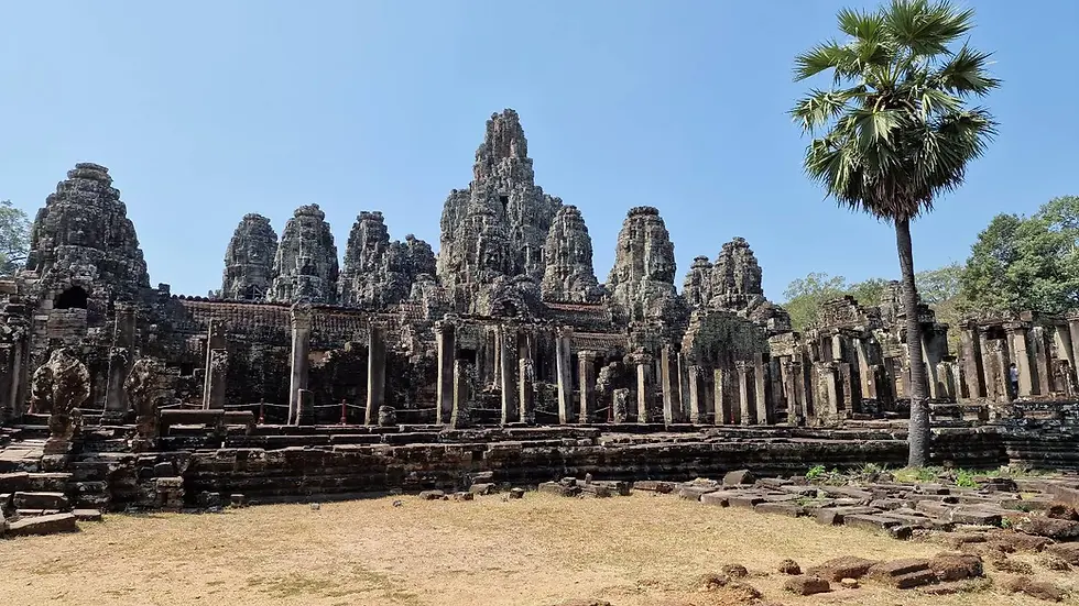 Ancient stone temple ruins with tall spires and columns under a clear blue sky. A lone palm tree stands nearby. Brown stone and greenery.