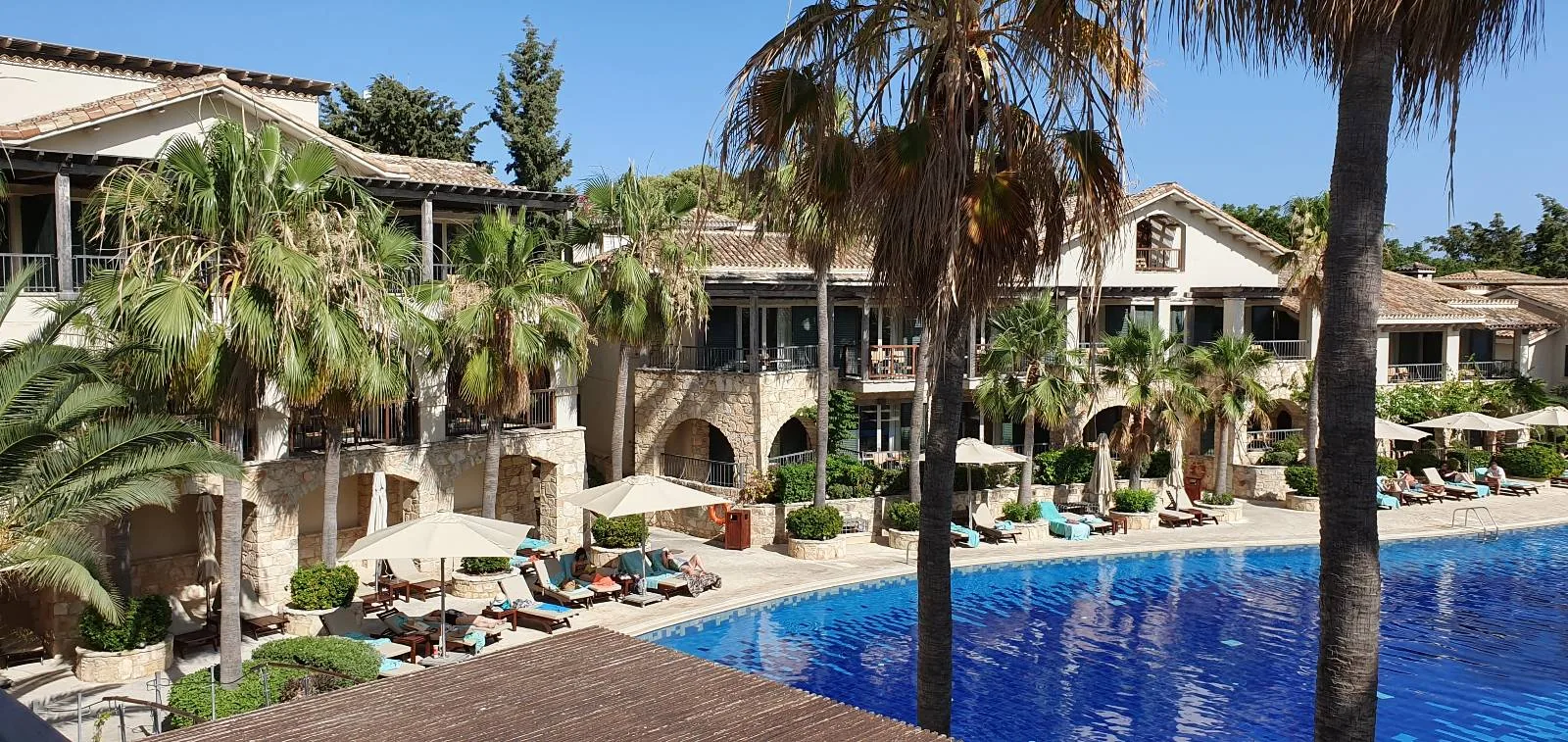 A luxurious outdoor pool area at a resort, surrounded by palm trees and elegant two-story buildings with arches and balconies. Sun loungers with parasols are arrayed along the pool's edge under a clear blue sky.