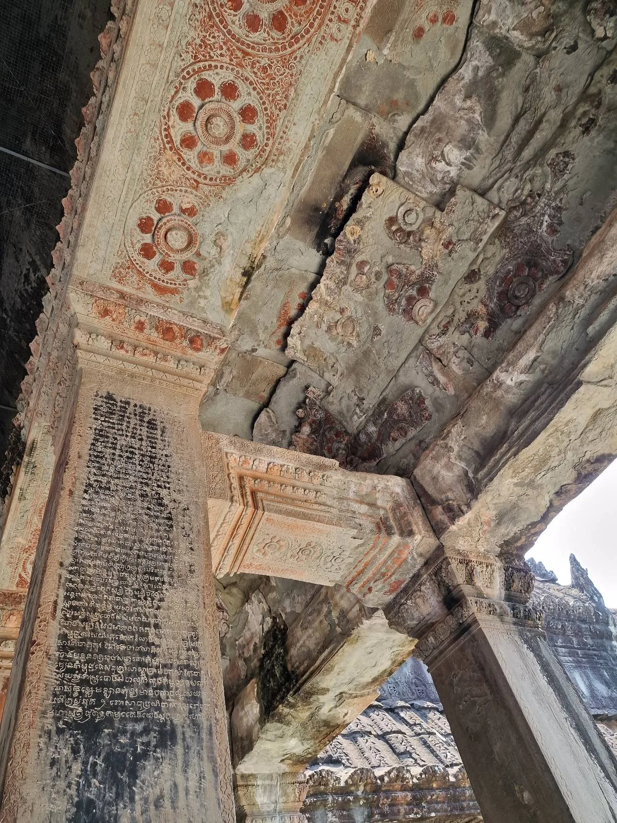 Ancient stone ceiling and pillars with faded red circular patterns and black script, showing cracks and weathering, likely part of a historic temple or ruin.