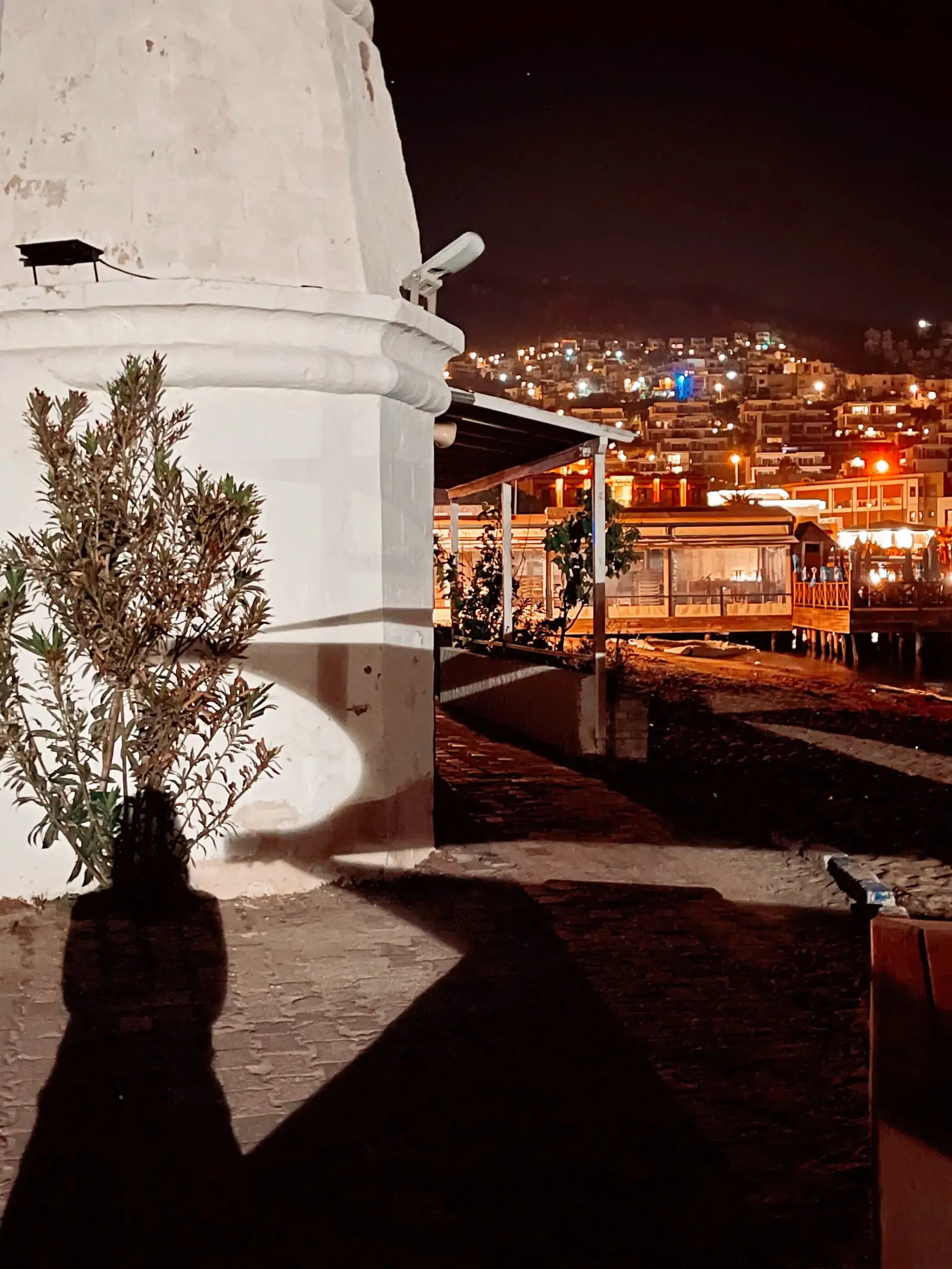 A night scene with a white structure, a small tree, and a person's shadow. In the background, a city is lit up, creating a vibrant display of lights on the hillside.