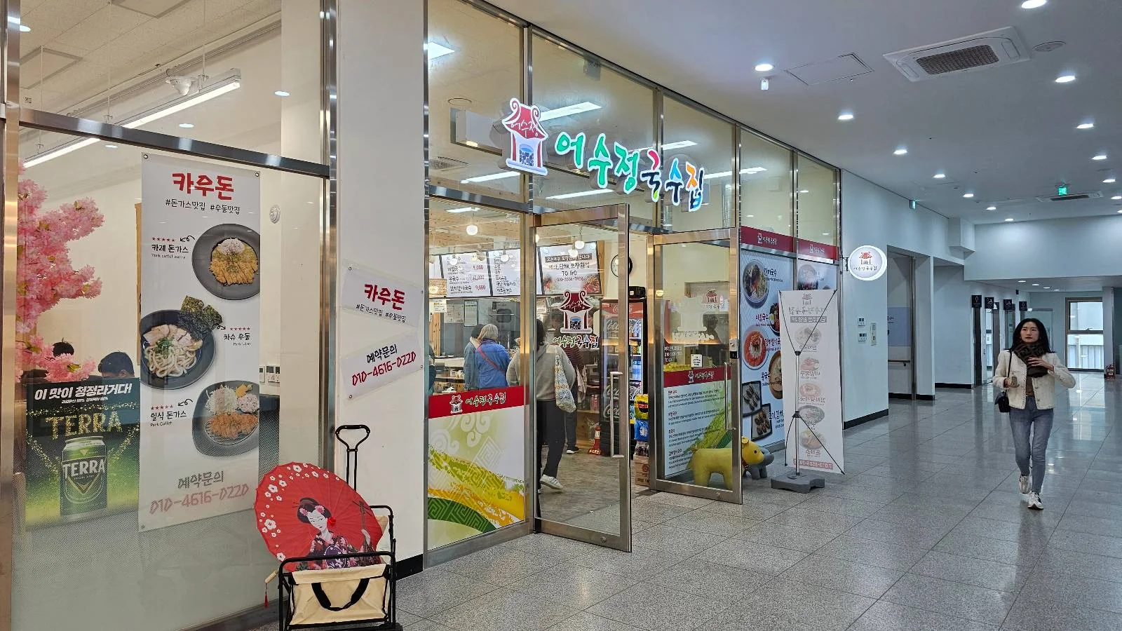 A Korean restaurant with glass walls and a brightly lit interior. There's a menu displayed on posters and a red suitcase with a cartoon character in front. A person walks by on the tiled floor.