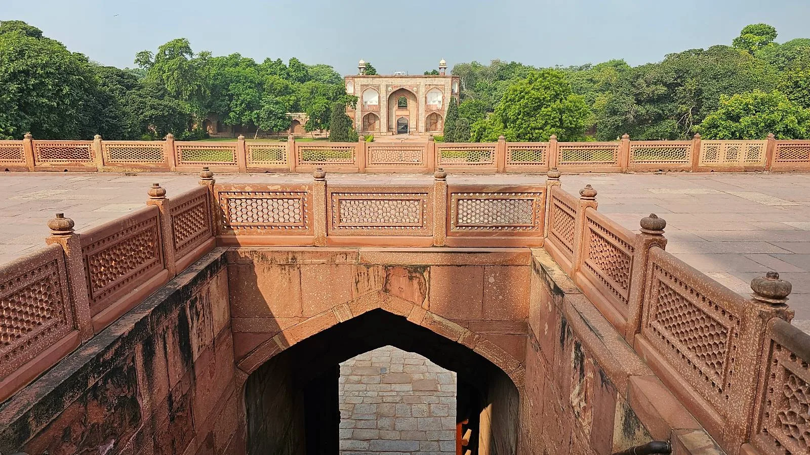 View of a historic sandstone structure with intricate carvings leading down to a staircase. In the distance, a grand building is surrounded by lush greenery under a clear sky.