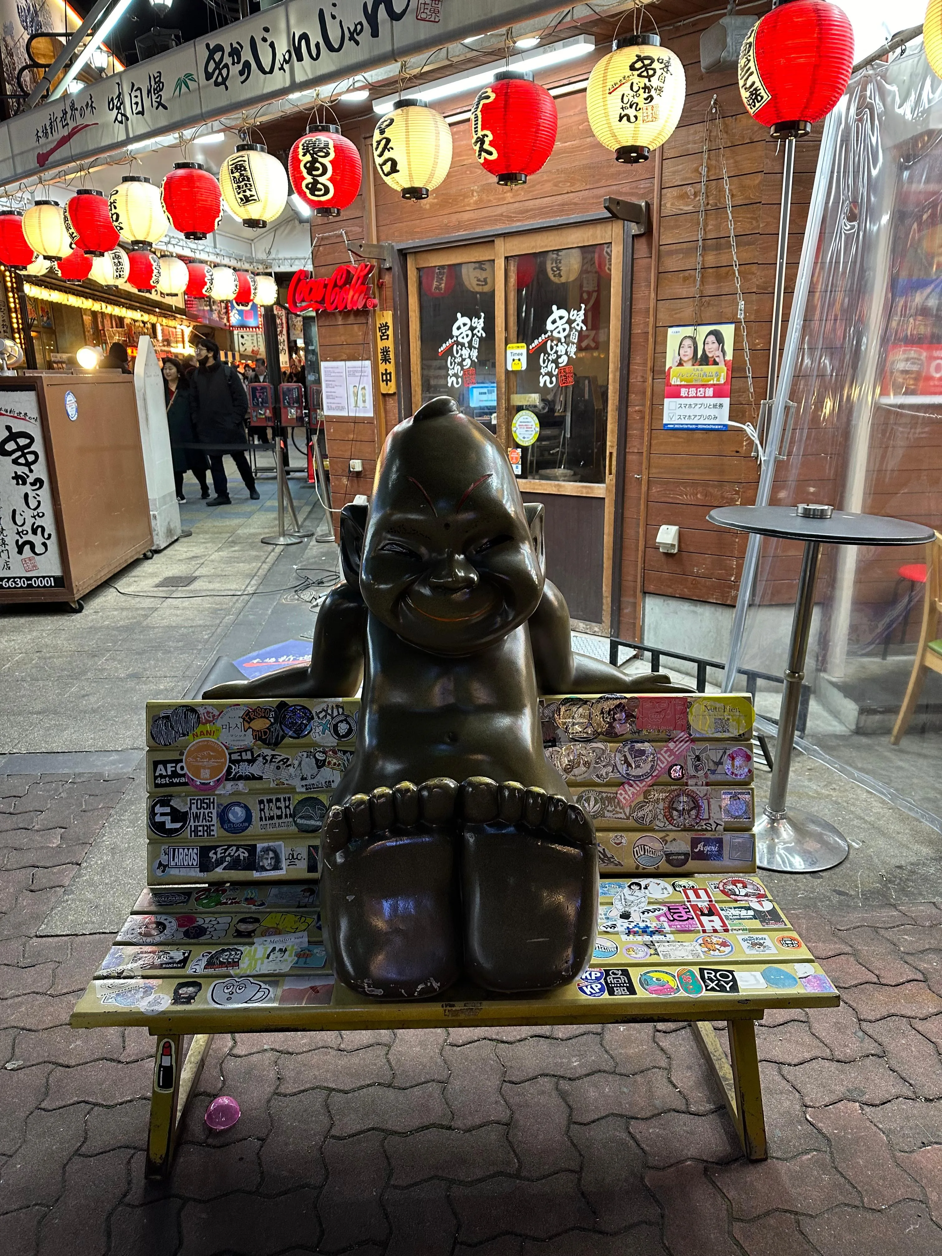 A bronze statue of a seated figure with large feet is displayed on a decorated bench covered in stickers, outside a restaurant adorned with red lanterns at night.