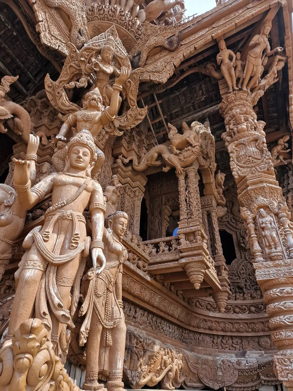 Intricately carved wooden statues and ornate columns at the Sanctuary of Truth in Thailand, showcasing detailed designs and mythical figures in a grand architectural setting.