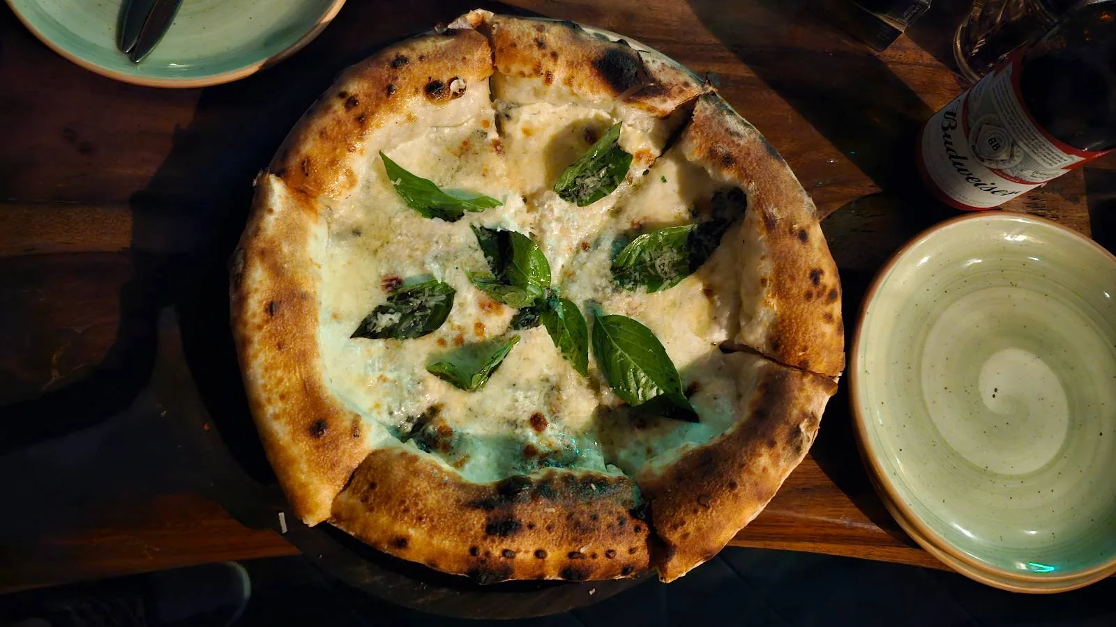 A freshly baked pizza with a golden-brown crust, topped with melted cheese and fresh basil leaves. It sits on a wooden table, surrounded by empty plates and silverware. The lighting is warm, highlighting the pizza's texture.