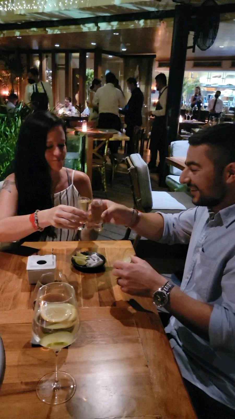 A woman and a man are seated at a wooden table in a dimly lit restaurant, clinking glasses. They appear to be enjoying a meal together. There is food and drinks on the table, with other diners and greenery visible in the background.