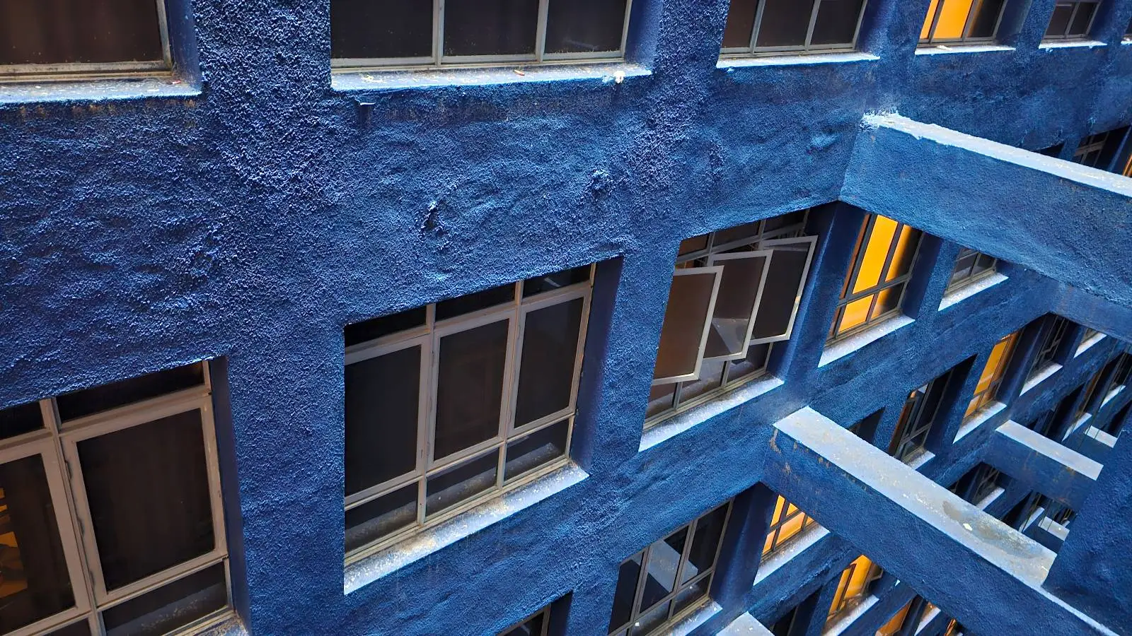 View of a blue-painted building exterior with multiple windows, some open, and illuminated yellow light visible from inside. The photo is taken at an angle, showing textured walls and architectural beams.