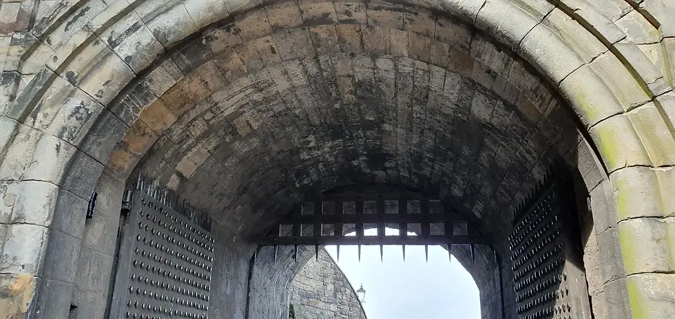 Stone archway with spiked metal gate partially open. Gray stones form textured ceiling. Background features a cloudy sky and a street lamp.