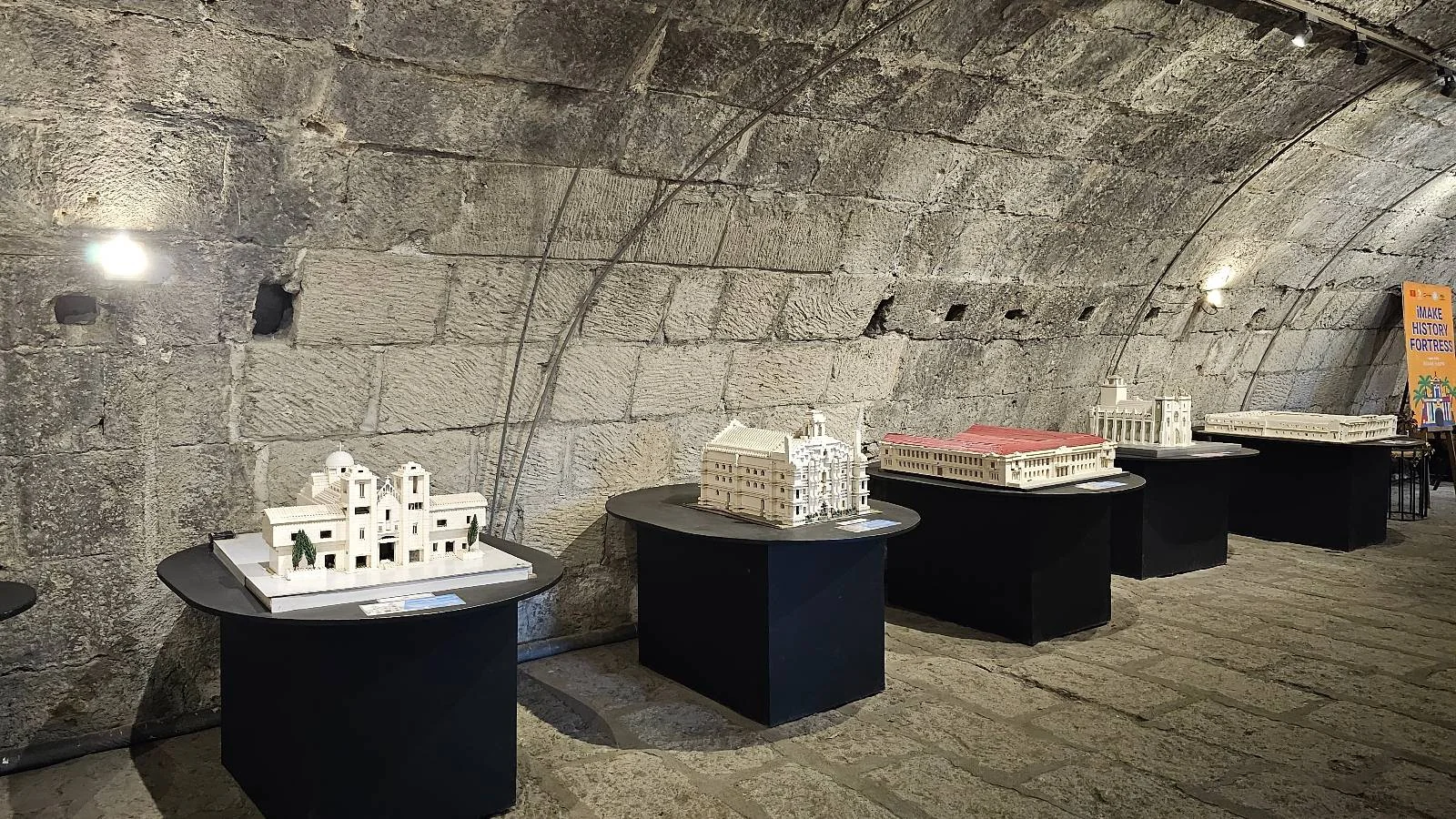A dimly lit stone-walled room displaying a series of architectural models on round black tables. Each model is detailed, showcasing different building designs. The arched ceiling adds a historic atmosphere to the exhibit.