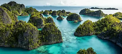 An aerial view of numerous small, jungle-covered limestone islands scattered throughout vibrant, clear turquoise lagoons under a bright sky.
