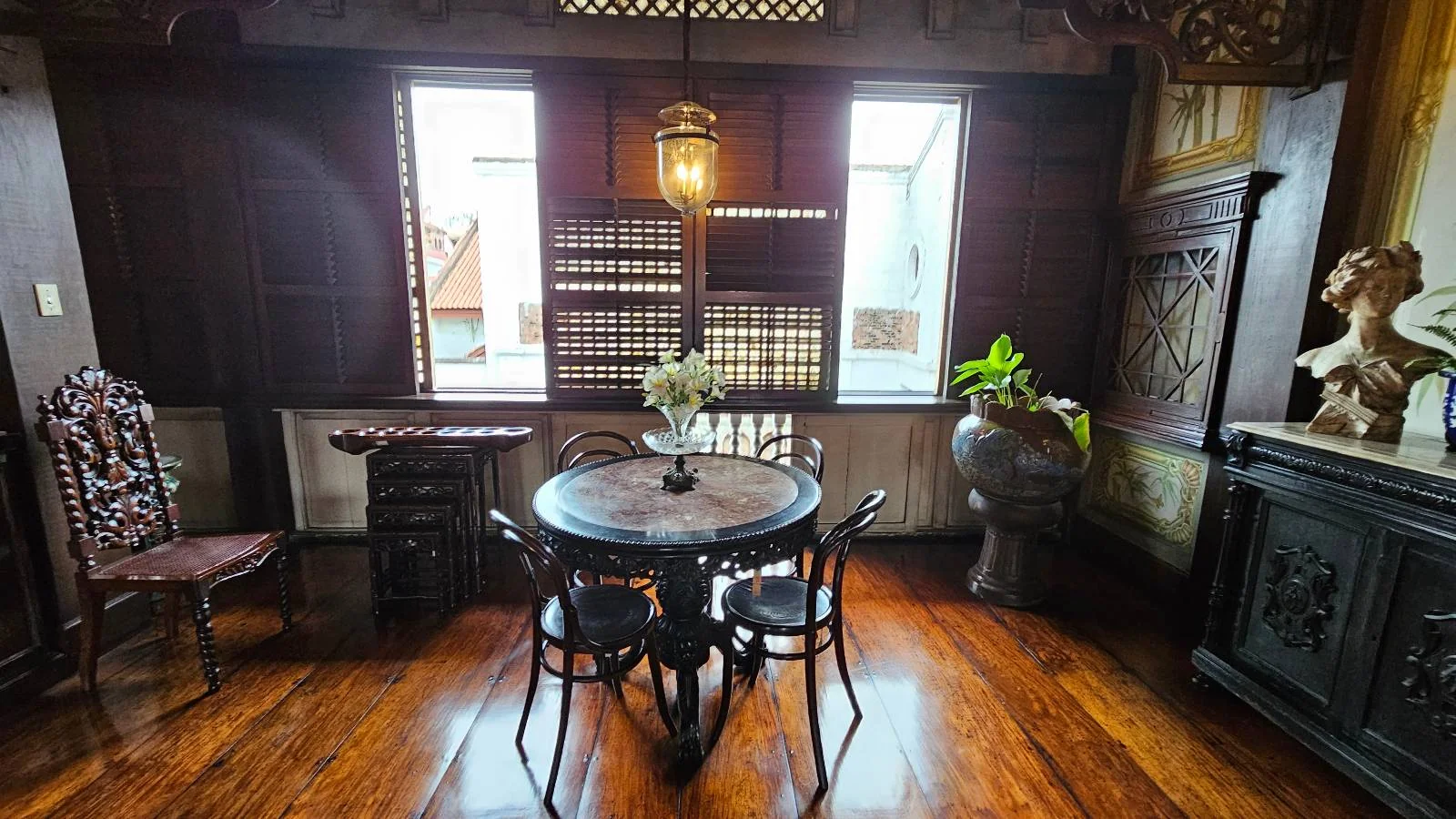 A cozy room with a round wooden table and four chairs. The floor is polished wood, and large windows let in natural light. Shelves with books line the walls, and a bust and plants add decor. A hanging light fixture illuminates the space.