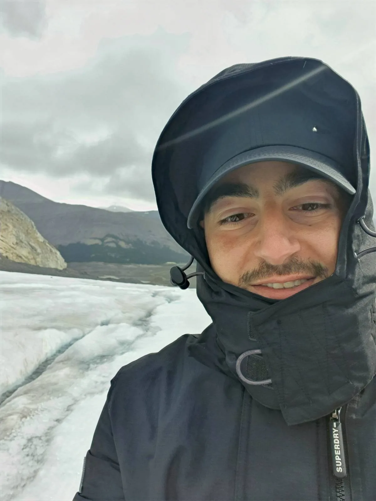 A person in a black jacket and hood smiles at the camera, standing on a snowy or icy landscape with mountains and a cloudy sky in the background.
