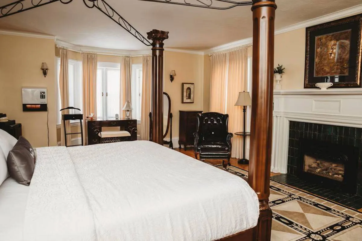 Elegant bedroom with four-poster bed, patterned floor, and fireplace. Cream curtains, dark armchair, desk by bay window, warm ambiance.