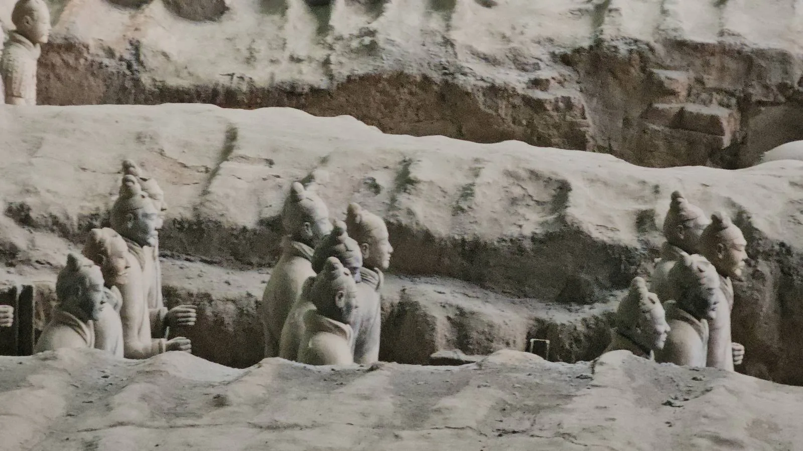 Partially buried ancient stone warrior statues with pointed helmets and detailed armor, set in an excavated, earth-covered archaeological site.