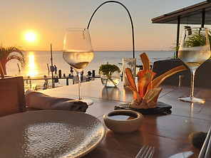 A table set for two with wine glasses and a dish of vegetable sticks beside a dipping sauce overlooks Mexico's stunning beaches at sunset. The glowing horizon and soft waves create a peaceful dining atmosphere.