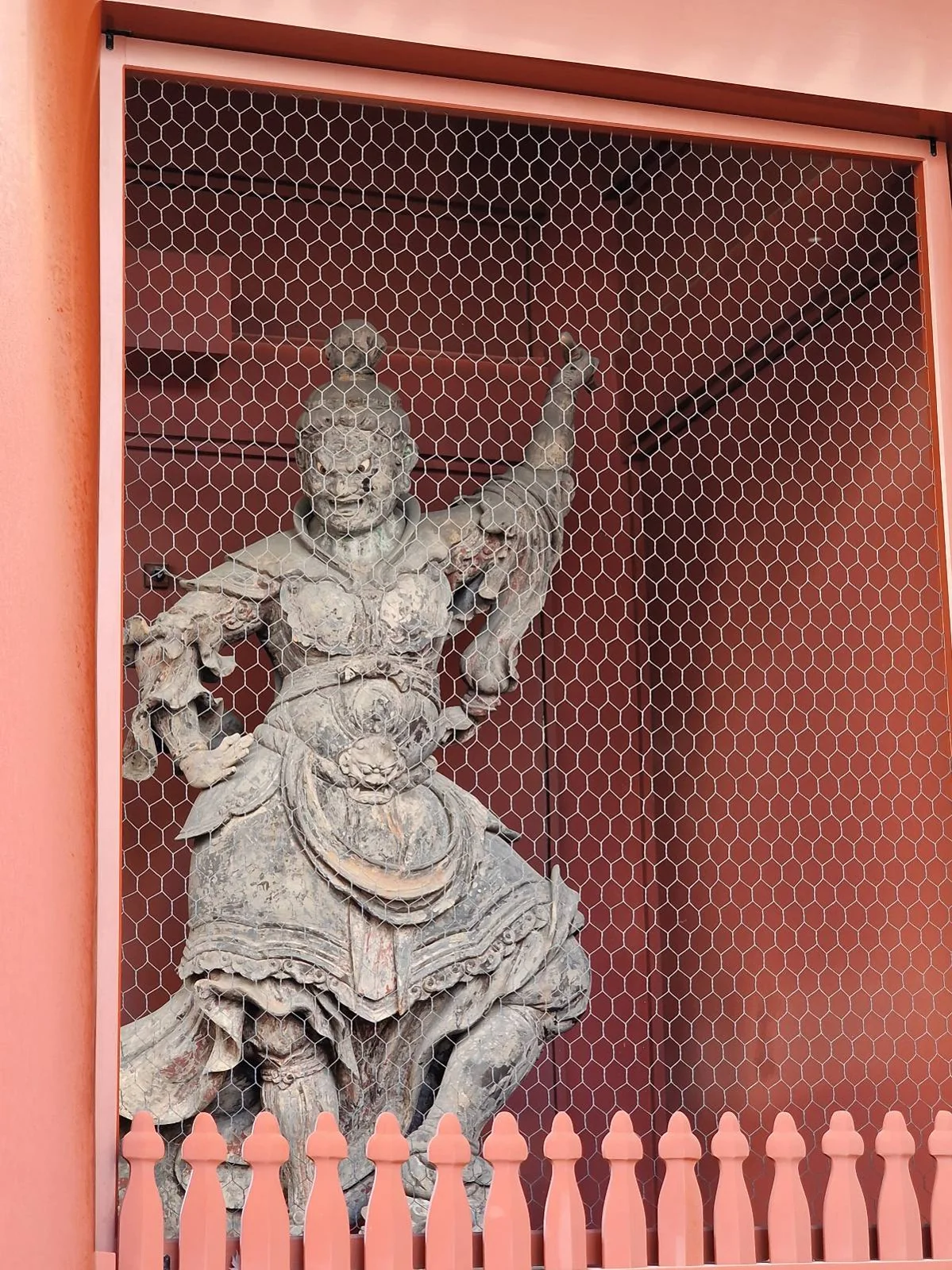 Stone warrior statue behind wire mesh, with armor details and dynamic pose. Set against red walls and framed by a red fence.