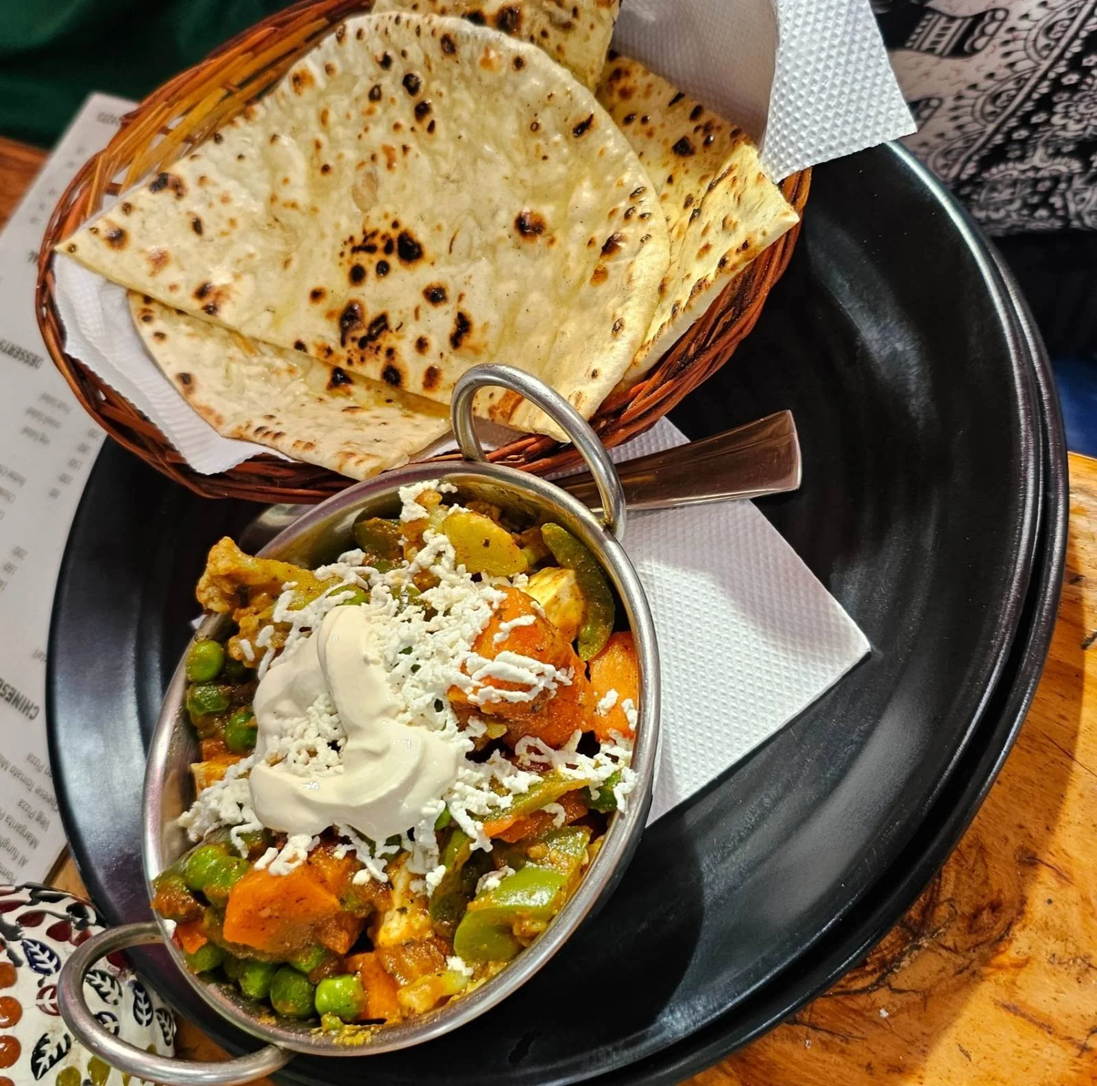 A bowl of mixed vegetable curry topped with cream and grated cheese is served with a basket of folded, grilled flatbreads on a black plate.A bowl of mixed vegetable curry topped with cream and grated cheese is served with a basket of folded, grilled flatbreads on a black plate.