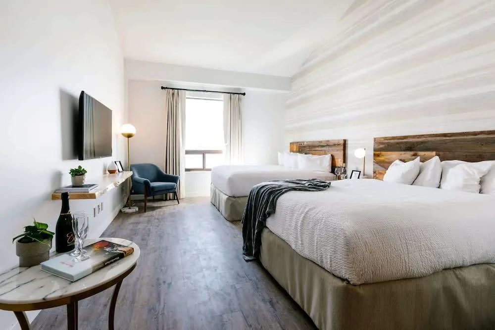 A hotel room with two large beds, each with white bedding and wooden headboards. A TV is mounted on the wall opposite the beds. There is a window with curtains, a blue chair, a small round table with a plant and bottle, and a shelf with decor.