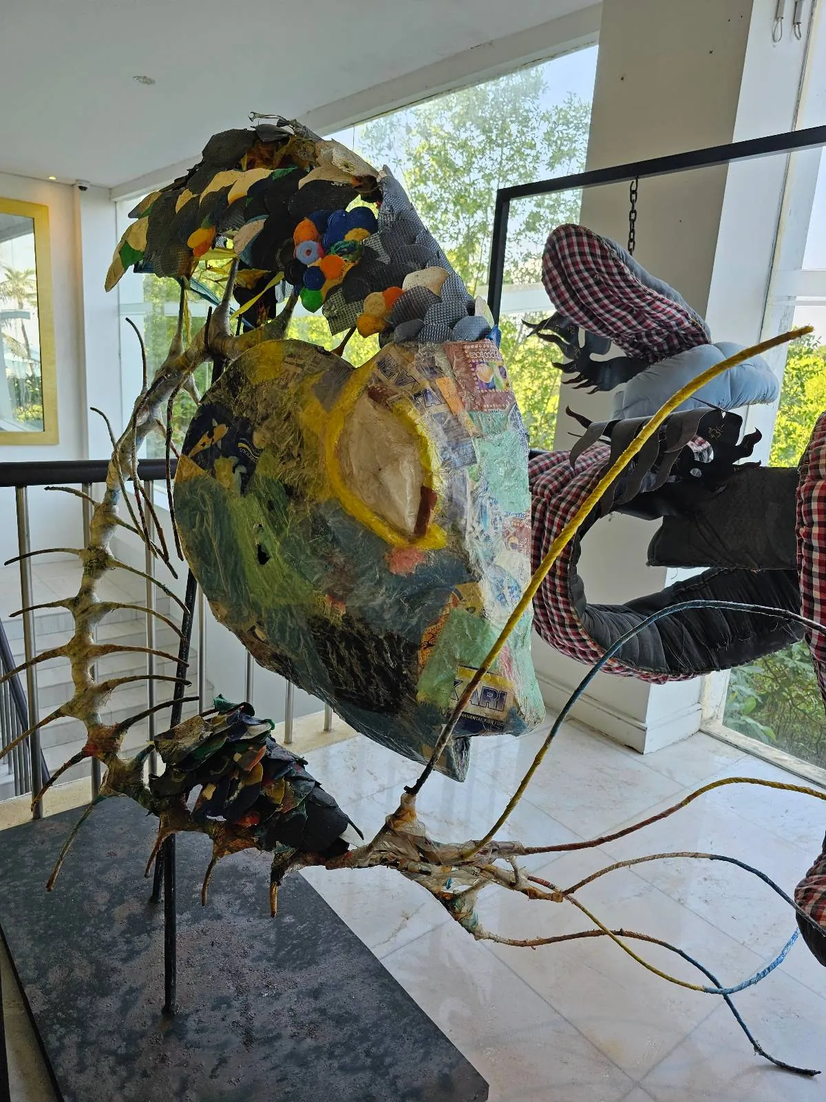 A colorful abstract sculpture made of mixed materials, featuring organic shapes and intertwined wires, is displayed indoors near large windows with greenery visible outside.