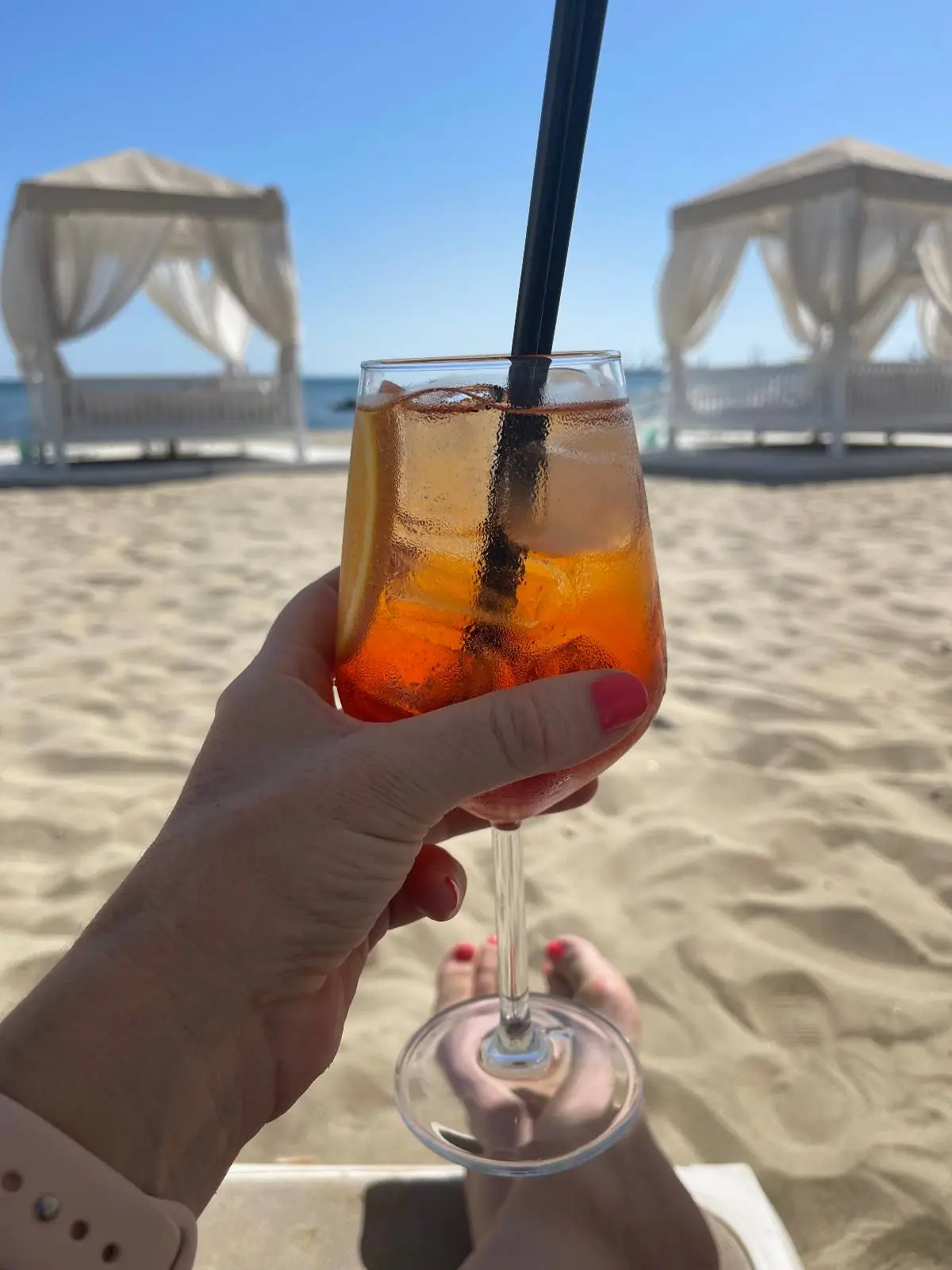 A hand holding a glass of orange cocktail with a black straw, on a sandy beach with white cabanas and blue sky in the background.