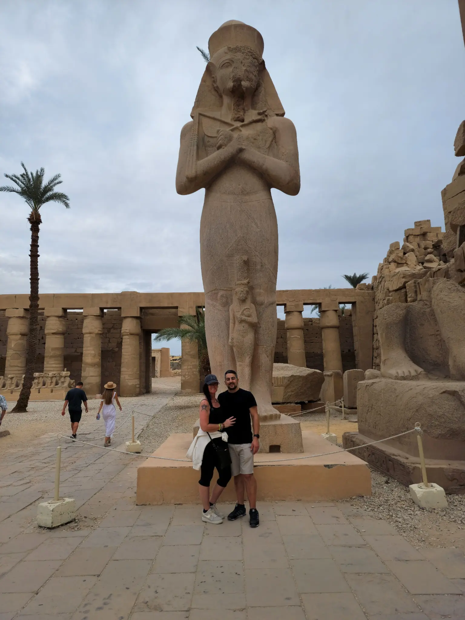 A large ancient statue stands in front of a temple structure with large columns. A man and a child pose in front of the statue. The sky is overcast, and a palm tree is visible on the left. Other tourists are in the background.