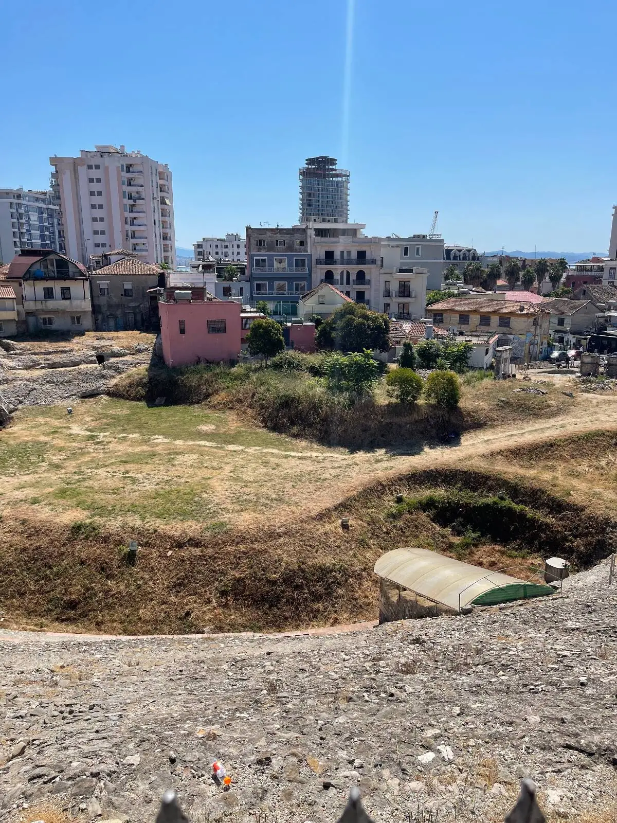 A construction site with patches of dry grass and dirt, surrounded by a mix of low-rise houses and taller modern buildings under a clear blue sky.