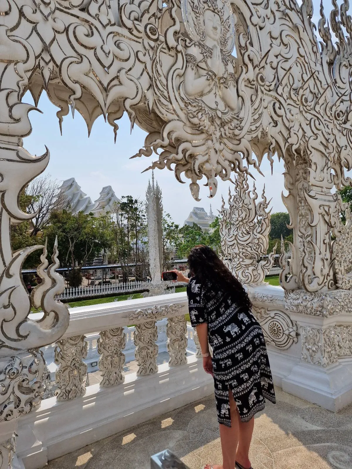 A person in a black and white patterned dress stands on a balcony adorned with intricate white carvings, looking towards an ornate garden with trees and sculptures under a clear sky.