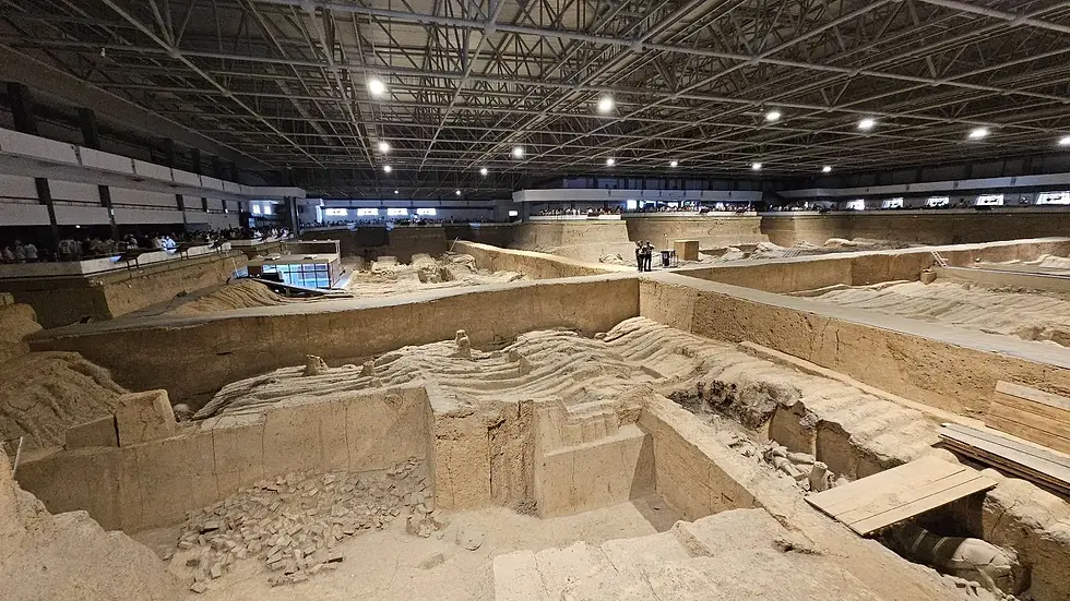 Vast indoor archaeological site with terracotta warrior excavations, dim lighting, metal ceiling beams, and people observing from the perimeter.