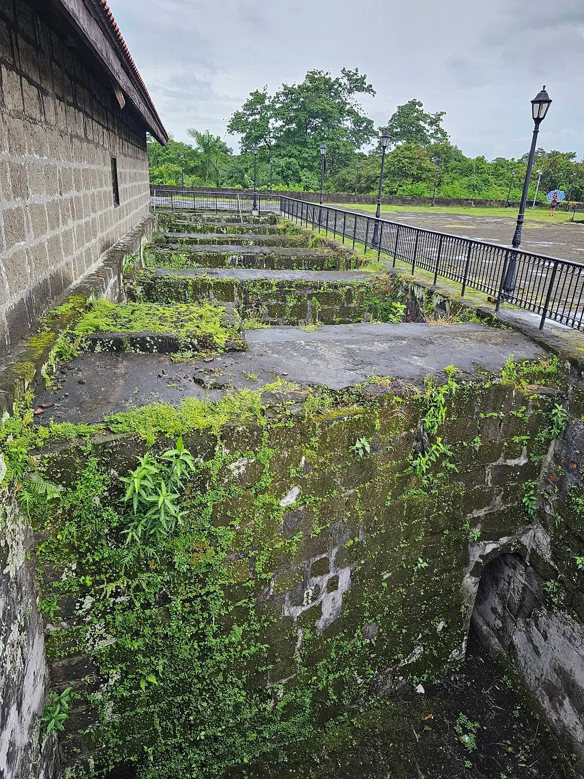View of an old stone structure with moss-covered walls and a green landscape in the background. The building has multiple rectangular sections and a black metal fence running alongside it. A tall lamppost stands near the fence under a cloudy sky.