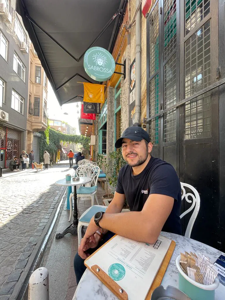 A man in a black t-shirt and cap sits at a small outdoor café table on a cobblestone street, smiling at the camera. There is a menu and a plate on the table, with buildings and more tables in the background.