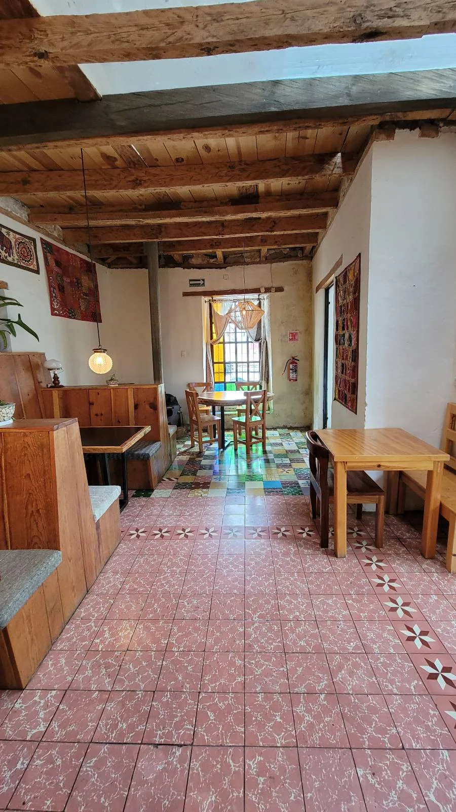 Cozy cafe interior with wooden booths and tables. The space features tiled pink and patterned floors, a stained glass window at the back, and a hanging pendant light. Wooden beams line the ceiling, creating a warm atmosphere.
