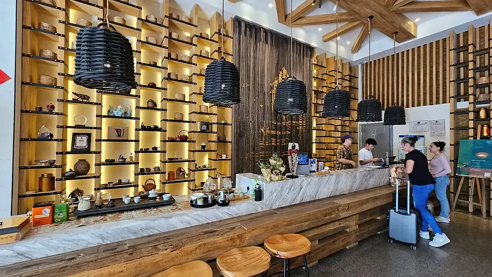 People at a wooden counter in a café with warm lighting. Shelves display teapots and items. Two people at the counter, two waiting with luggage.