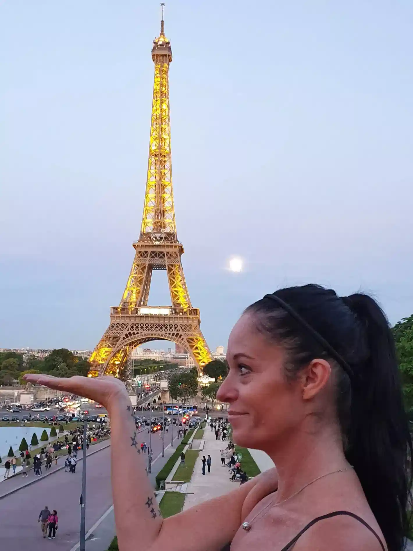A woman stands outdoors, holding her hand up to make it appear as if she is supporting the illuminated Eiffel Tower in the background at dusk.