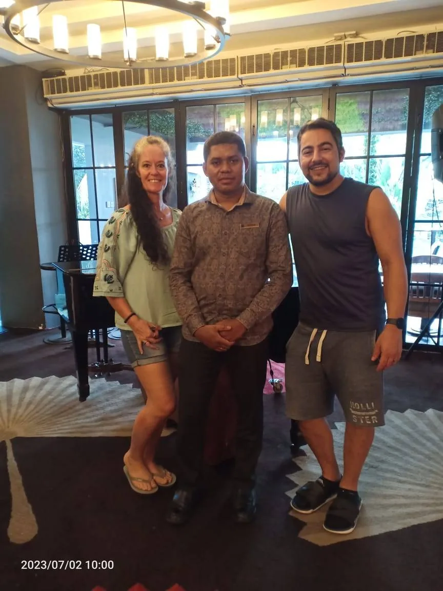 A man and a woman stand on either side of a person wearing a patterned shirt and dark pants in a well-lit room with a piano and chandelier in the background. The setting appears casual and relaxed.