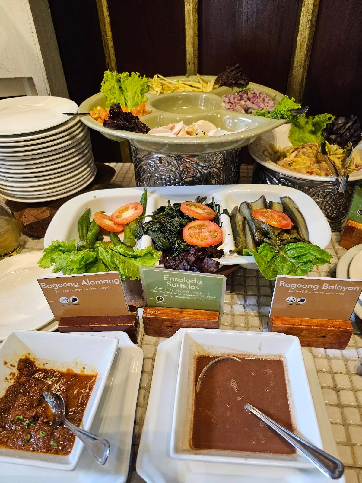 A buffet setup with plates of salad greens, sliced tomatoes, whole vegetables, and two brown sauces in white square dishes. Stacks of plates are on the side.
