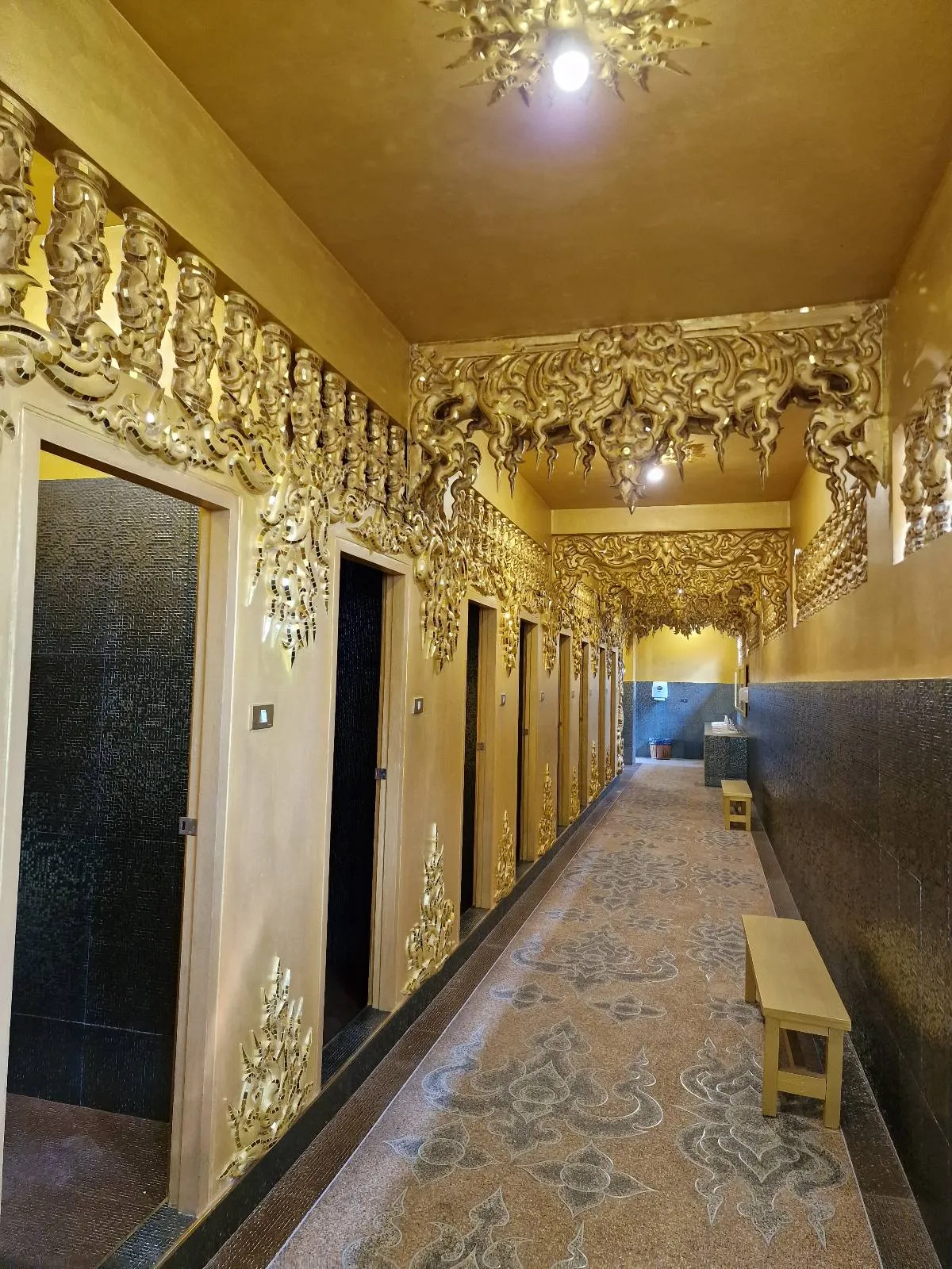 A lavish hallway with ornate golden decorations on the ceiling and walls. The passage is lined with several wooden doors, and a patterned carpet covers the floor. A wooden bench is placed along the wall. The lighting casts a warm glow.