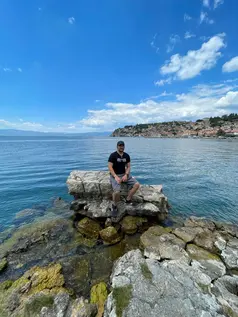 A person sits on a large rock formation at the edge of Lake Ohrid, one of the oldest lakes in Europe. A town can be seen in the distance under a partly cloudy sky, with more rocks scattered around the tranquil shoreline.