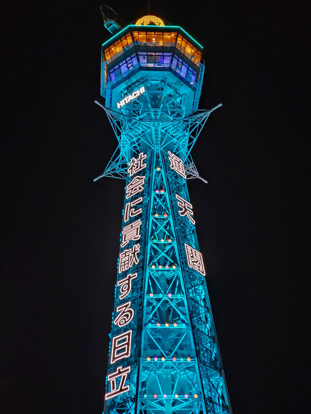 A tall observation tower brightly illuminated in blue and decorated with colorful lights, viewed from the base looking up against a dark night sky.