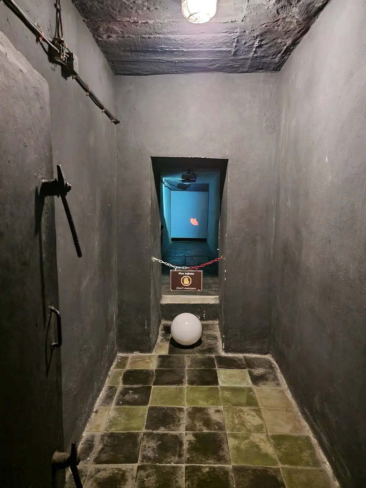 A narrow, dimly lit room with stone walls and a tiled floor. A small door with a caution sign and red tape blocks access. A white sphere is placed on the floor in the center. A single light bulb hangs from the ceiling.
