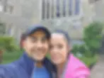 A couple smiling in front of a historic stone building with large windows. One wears a pink jacket, the other a blue cap. Lush greenery surrounds.