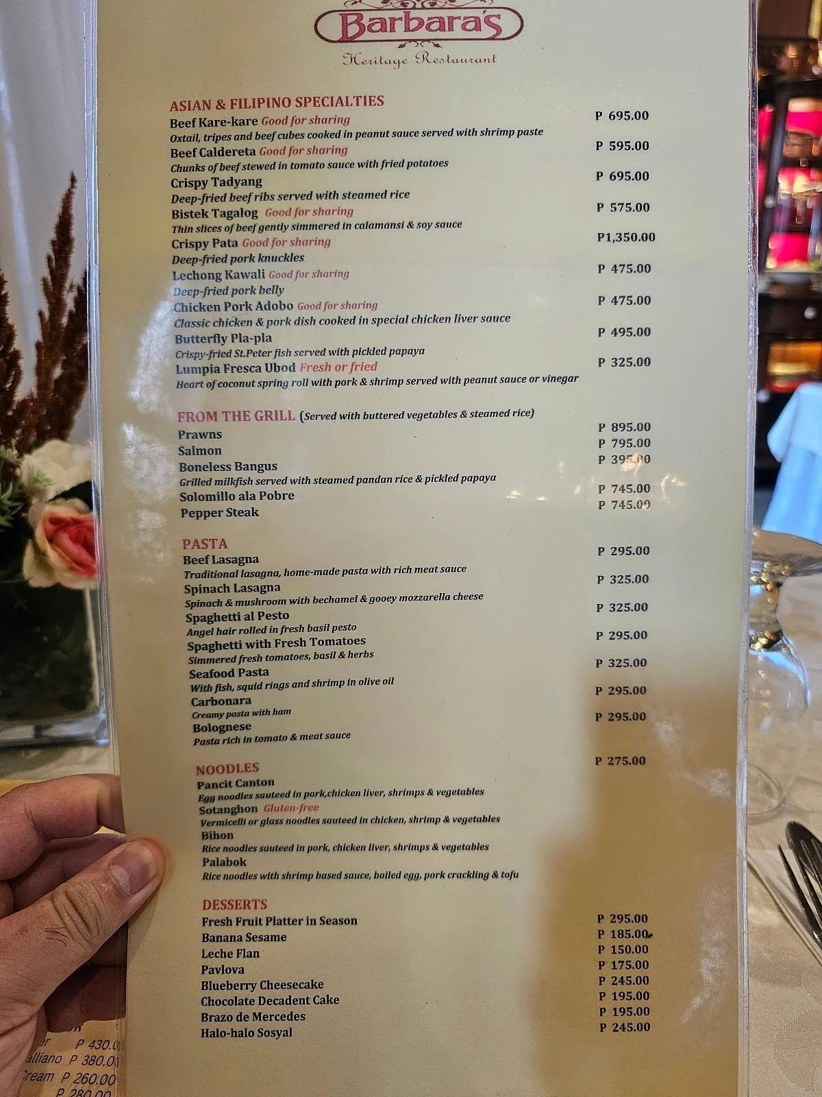 A hand holding a restaurant menu featuring appetizers, mains, pasta, desserts, beverages, and prices listed in Philippine pesos.