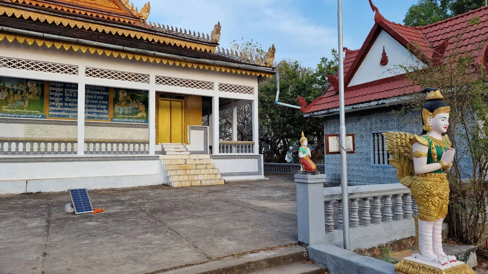 A traditional Thai temple with ornate golden and white details, yellow doors, and a statue of a figure with angel wings and a gold costume at the entrance. Another smaller building is visible on the right.