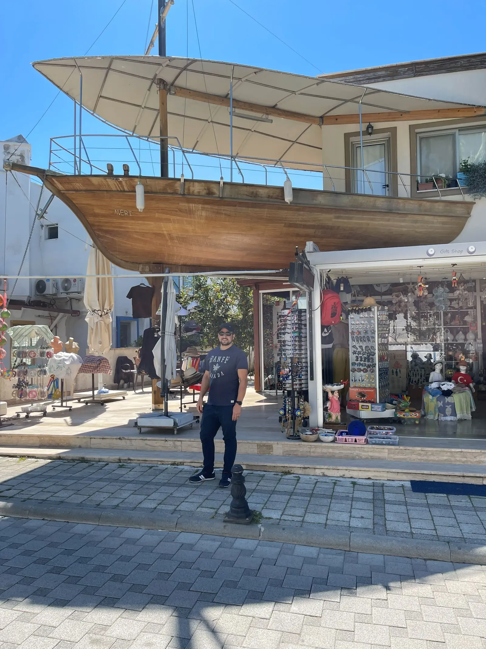 A man stands on a cobblestone street in front of a building with a large wooden boat structure mounted above its entrance, under a clear blue sky.