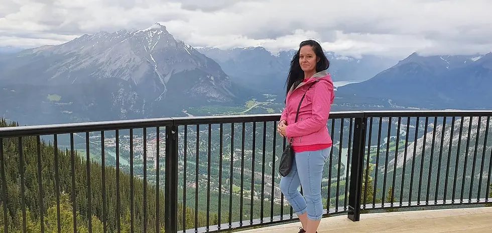 A person in a pink jacket stands by a railing with a mountainous landscape in the background. Cloudy sky above.