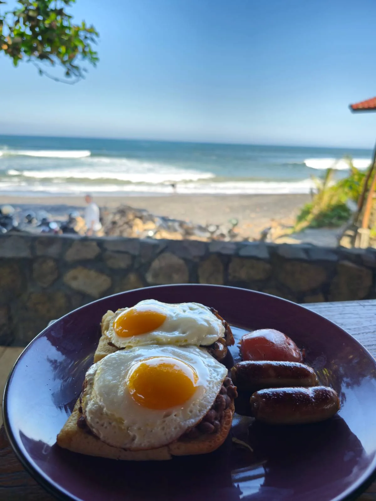 A plate with two sunny-side-up eggs on toast, accompanied by sausages, with a scenic ocean view in the background. Waves crash against the shore under a clear blue sky, framed by a tree and part of a building.