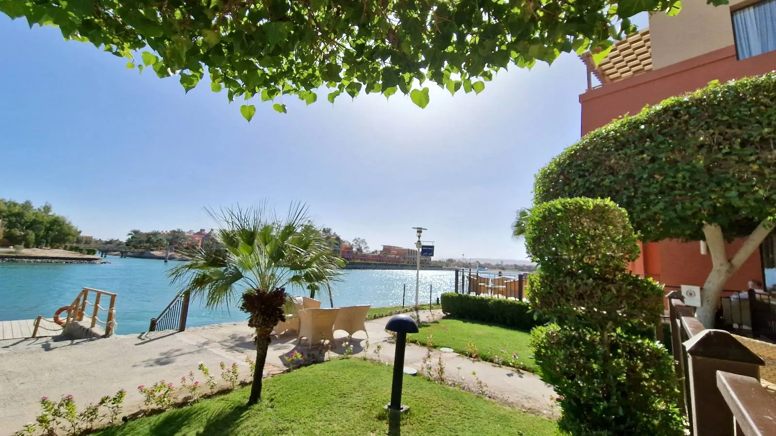 A waterside patio with green shrubs, palm trees, and outdoor seating next to a calm blue water body under a clear sky.